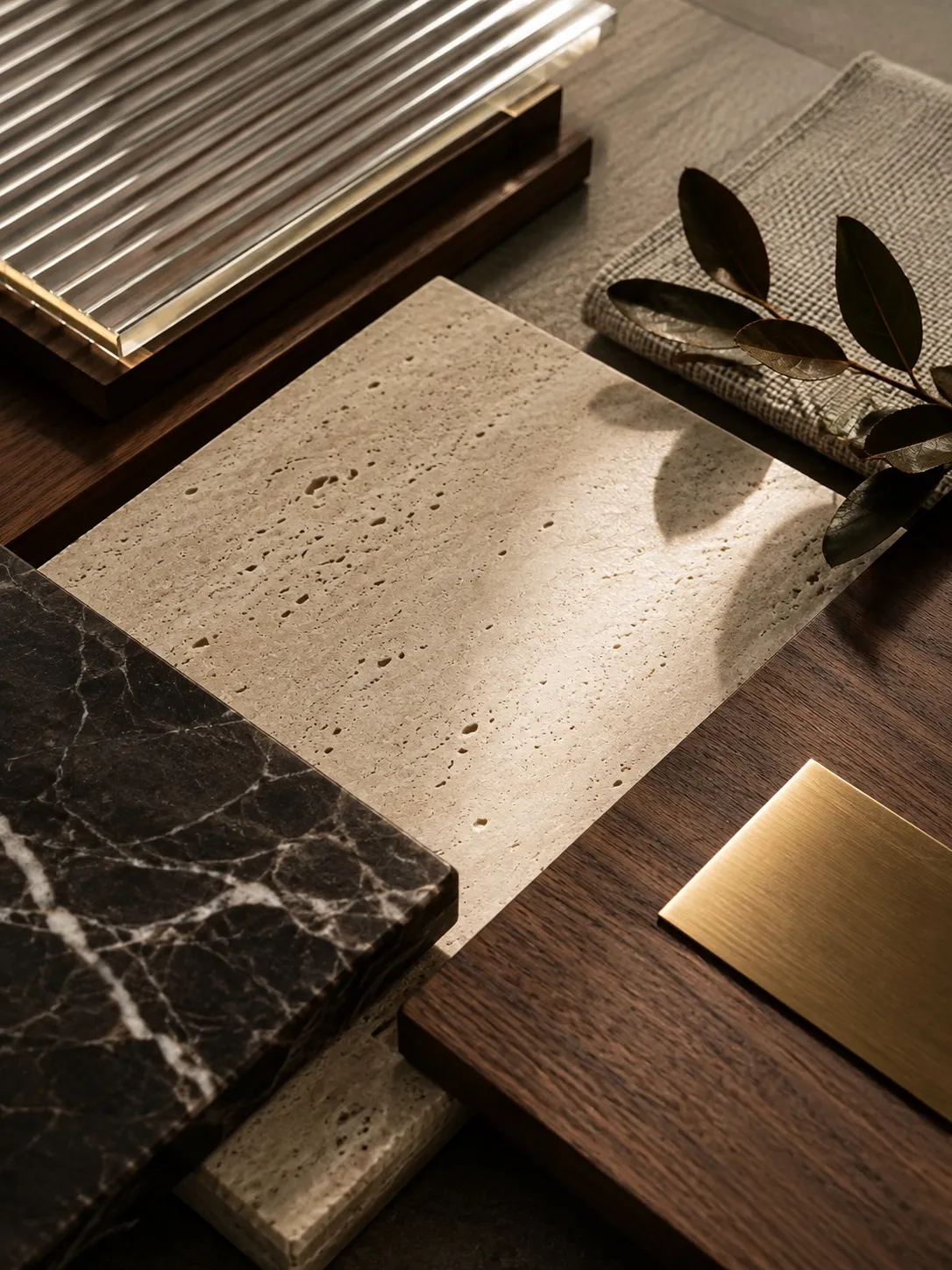 Material samples laid out on walnut in raking light — a travertine slab, dark veined marble, a brushed brass sheet, fluted glass and linen